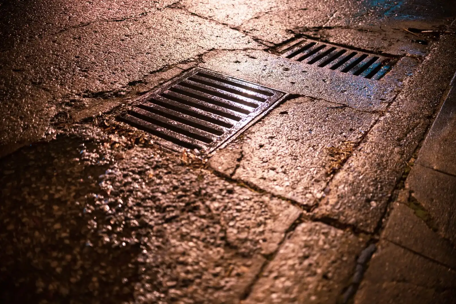 Deux grilles d'avaloir en fonte encastrées dans un pavage mouillé, filet d'eau ruisselant vers elles