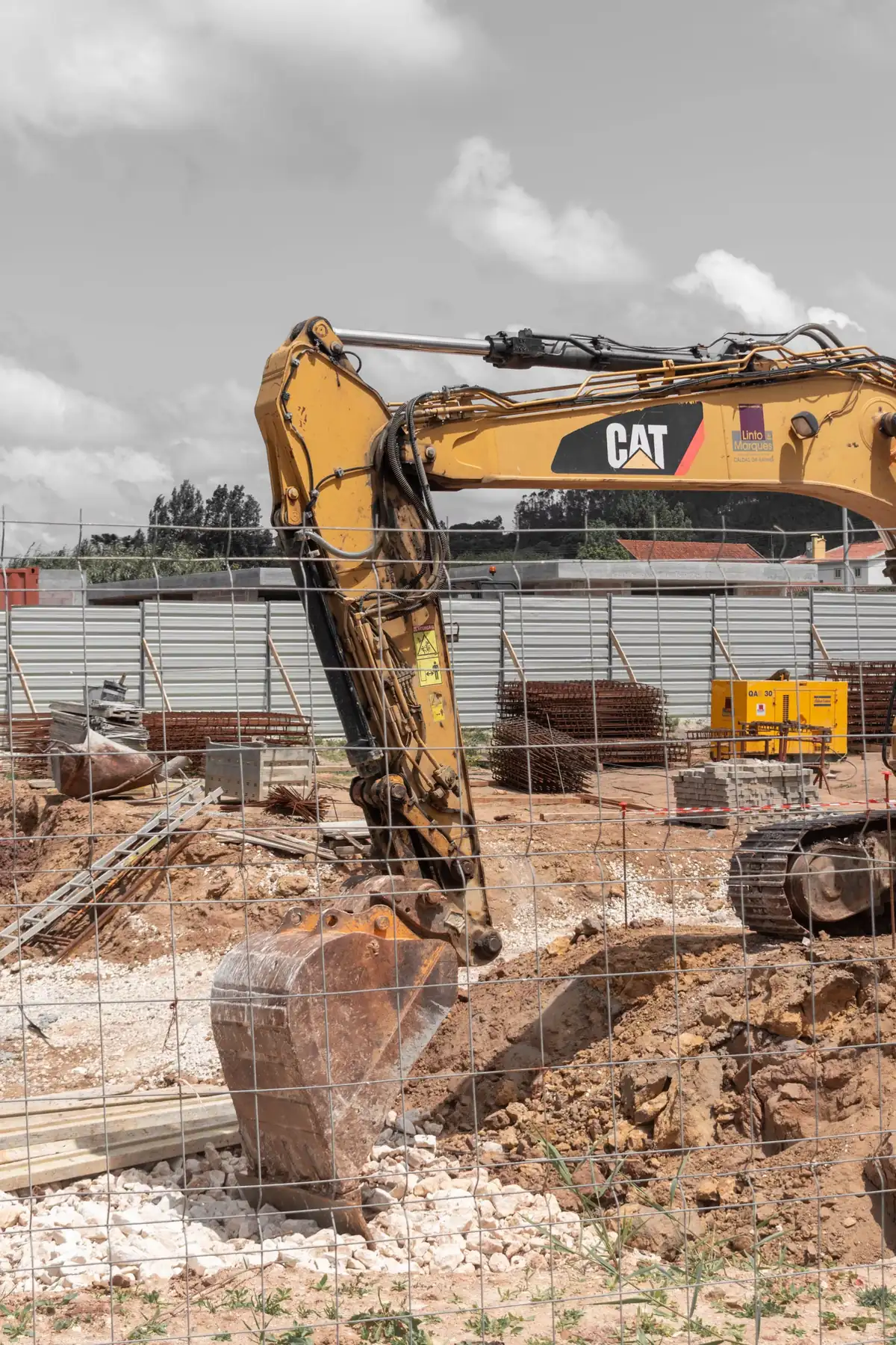 Godet d'une pelle mécanique posé au fond d'une fouille de fondation, aciers et matériaux stockés derrière la clôture de chantier